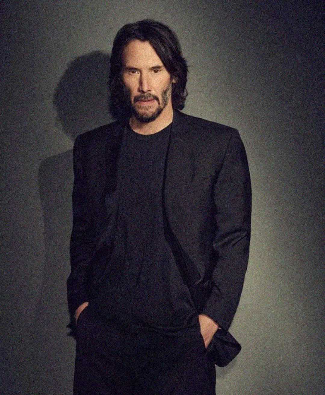 A man with shoulder-length dark hair and a beard stands against a plain backdrop, wearing a black suit and black shirt with his hands in his pockets, looking confidently at the camera.