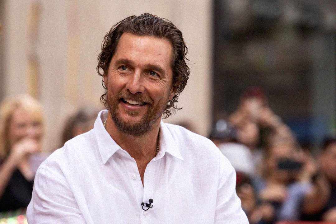 A man with wavy brown hair and a beard, wearing a white button-up shirt, smiles while sitting outdoors. He has a microphone clipped to his shirt, and blurred people and buildings are in the background.