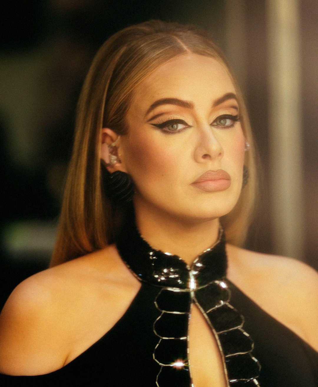 A woman with long straight hair, dramatic winged eyeliner, and nude lipstick gazes forward. She wears a black halter dress with shiny details, and the background is softly blurred.