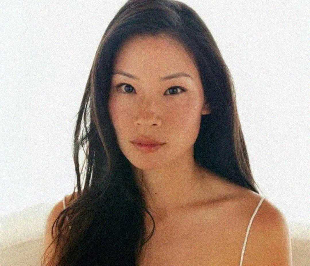 A woman with long dark hair and fair skin gazes at the camera. She is wearing a thin-strap top and is set against a soft, light background, giving the image a calm and serene feel.