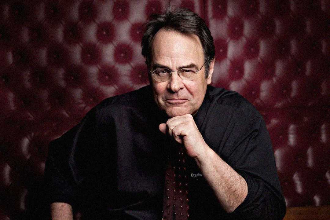 A middle-aged man with glasses and dark hair sits in front of a tufted red leather background, resting his chin on his hand and wearing a dark shirt and patterned tie.