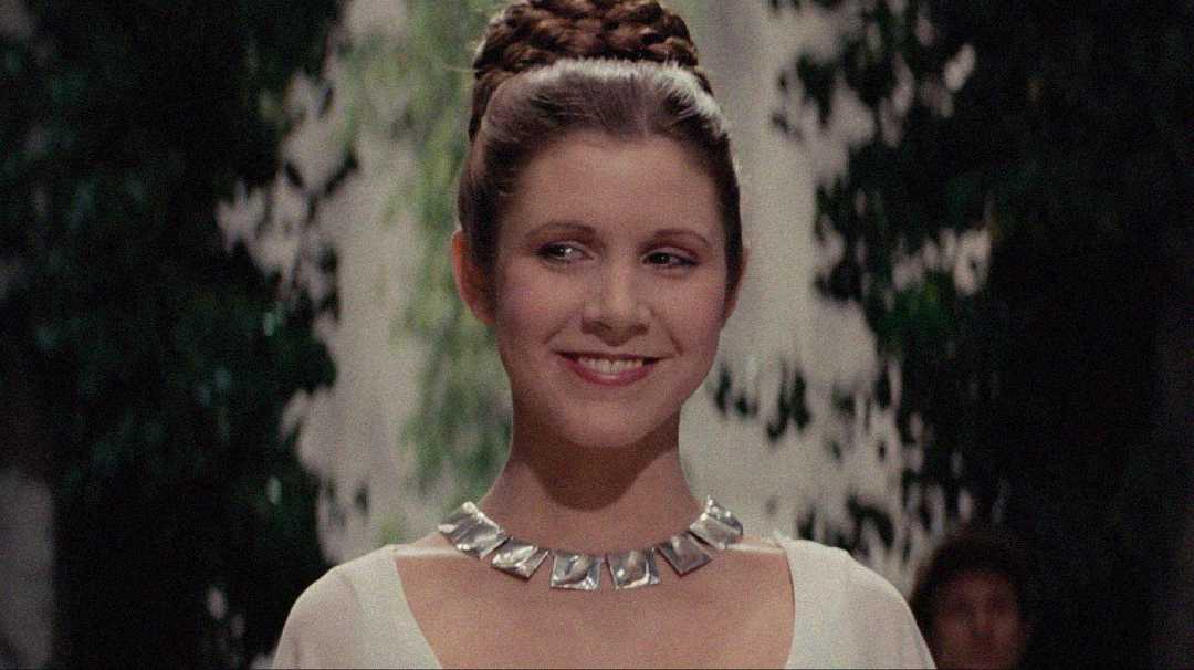 A woman with braided brown hair and a silver geometric necklace smiles while standing outdoors, with greenery and blurred people in the background.