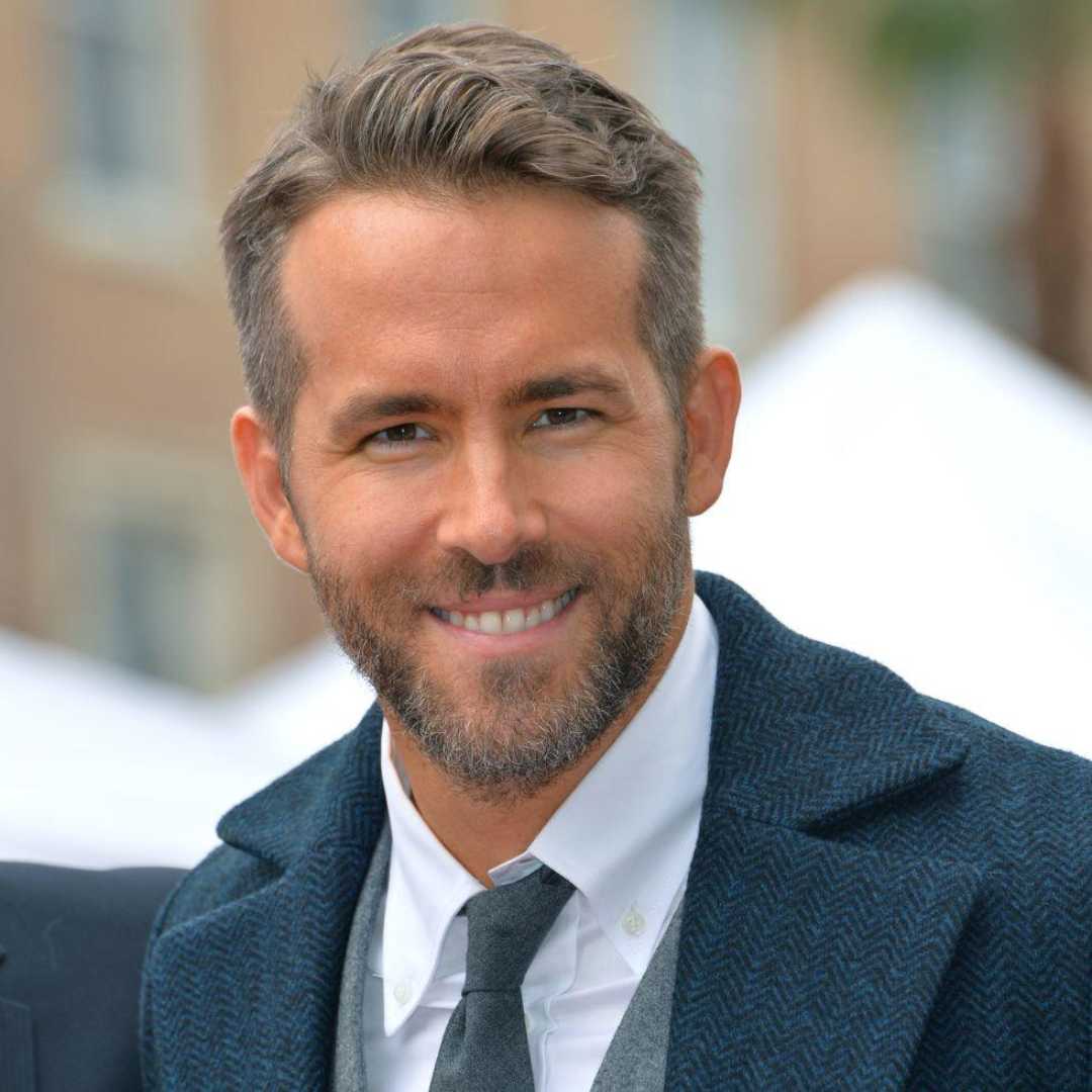 A man with short brown hair and a beard smiles at the camera. He is wearing a blue textured coat over a suit and tie. The background is blurred, showing an outdoor setting with a building and some greenery.