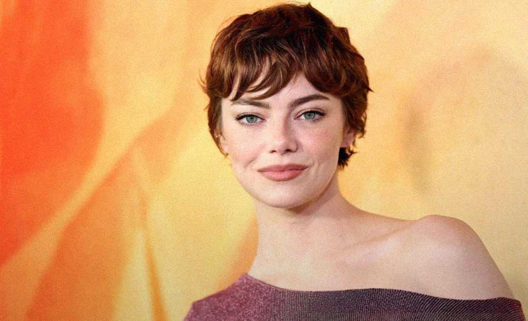 A woman with short brown hair and blue eyes is smiling softly at the camera. She wears a purple off-the-shoulder top and stands in front of a yellow-orange gradient background.