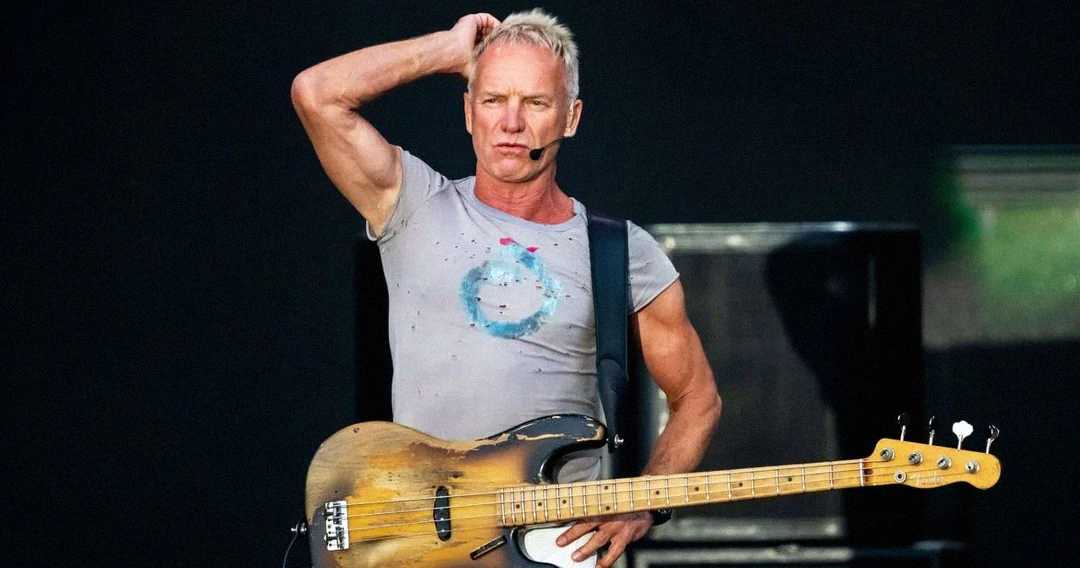 A musician with short blond hair stands on stage holding a bass guitar, wearing a headset microphone and a fitted gray t-shirt. One hand is on his hip and the other rests on his head. The background is dark.