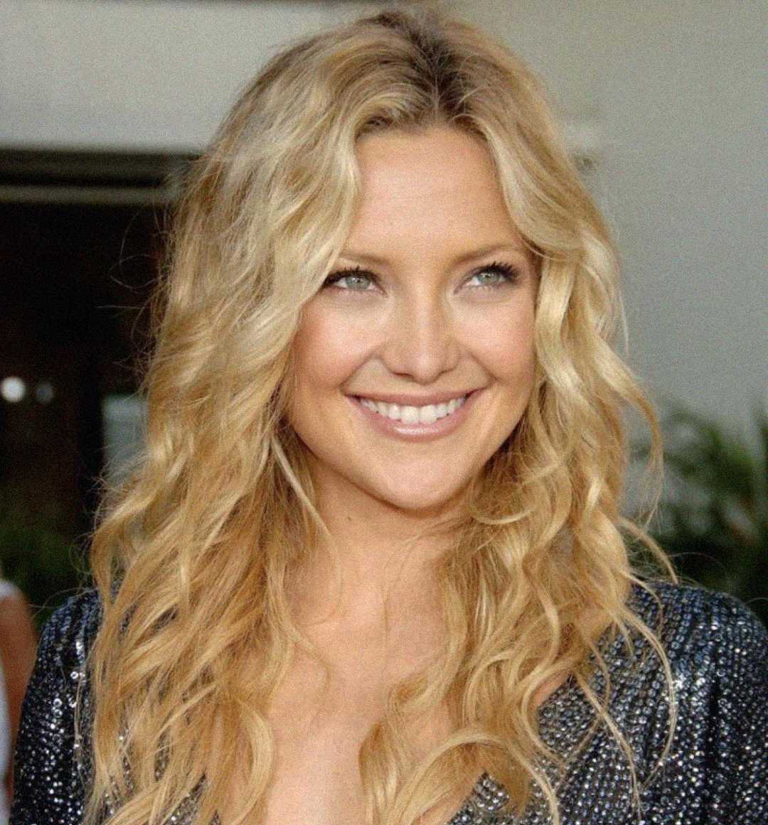 A woman with long, wavy blonde hair is smiling. She is wearing a dark, sparkly top and is standing in a well-lit setting with plants and blurred background elements.