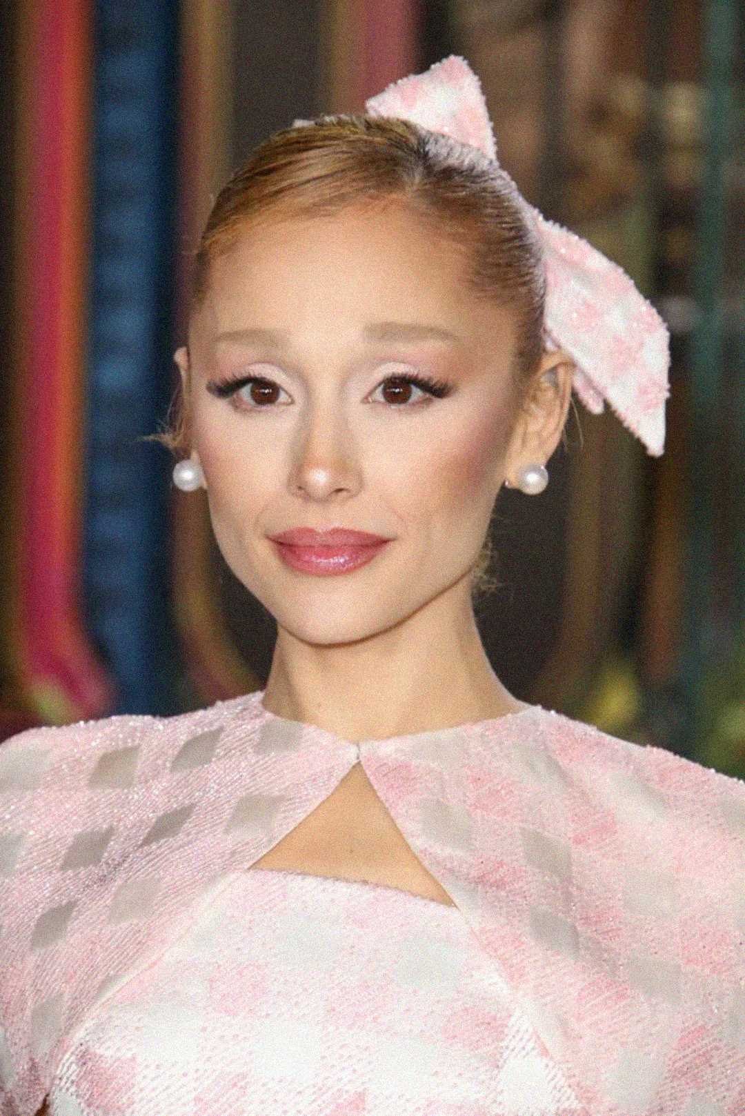 A woman with fair skin and light brown hair in a sleek updo, wearing a large pink bow, pearl earrings, and a pink checkered outfit, smiles softly against a blurred colorful background.