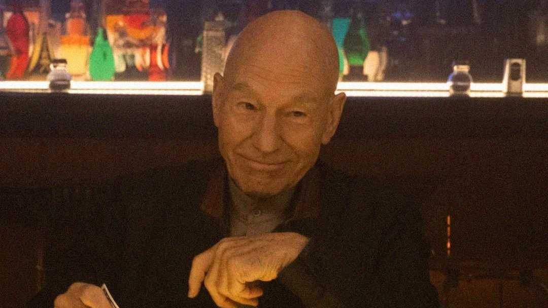 An older man with a bald head sits at a dimly lit bar, smiling subtly while holding something in his hand. Colorful bottles are visible on a lit shelf behind him.