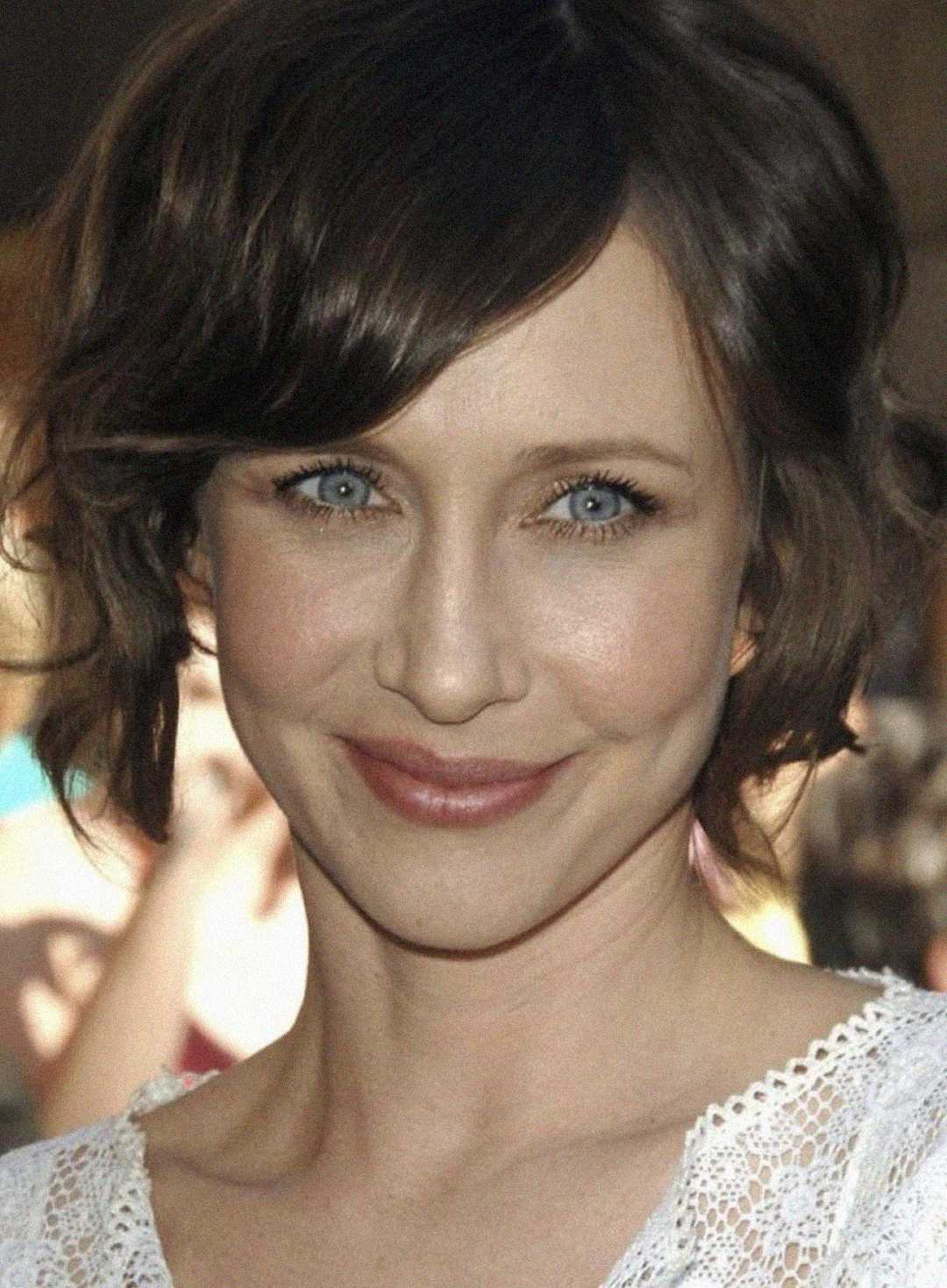 A woman with short brown hair, blue eyes, and light makeup smiles softly at the camera. She is wearing a white lace top and is standing in front of a blurred outdoor background.