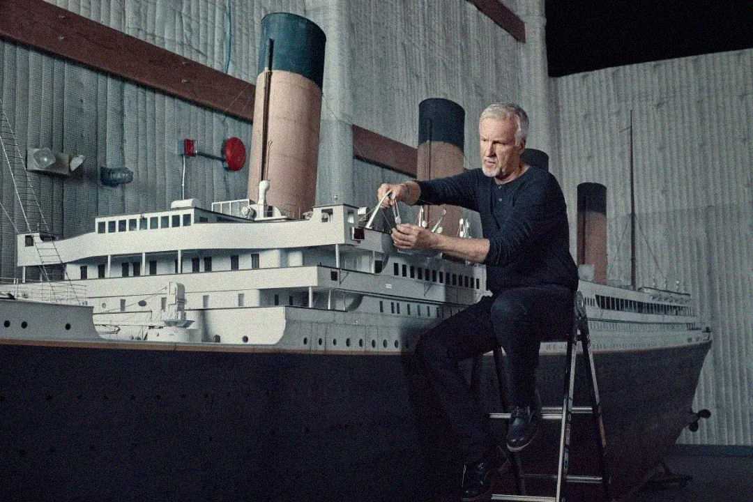 A man sits on a stepladder, working on a large, detailed model of an ocean liner ship inside a spacious, industrial-looking room with corrugated metal walls.