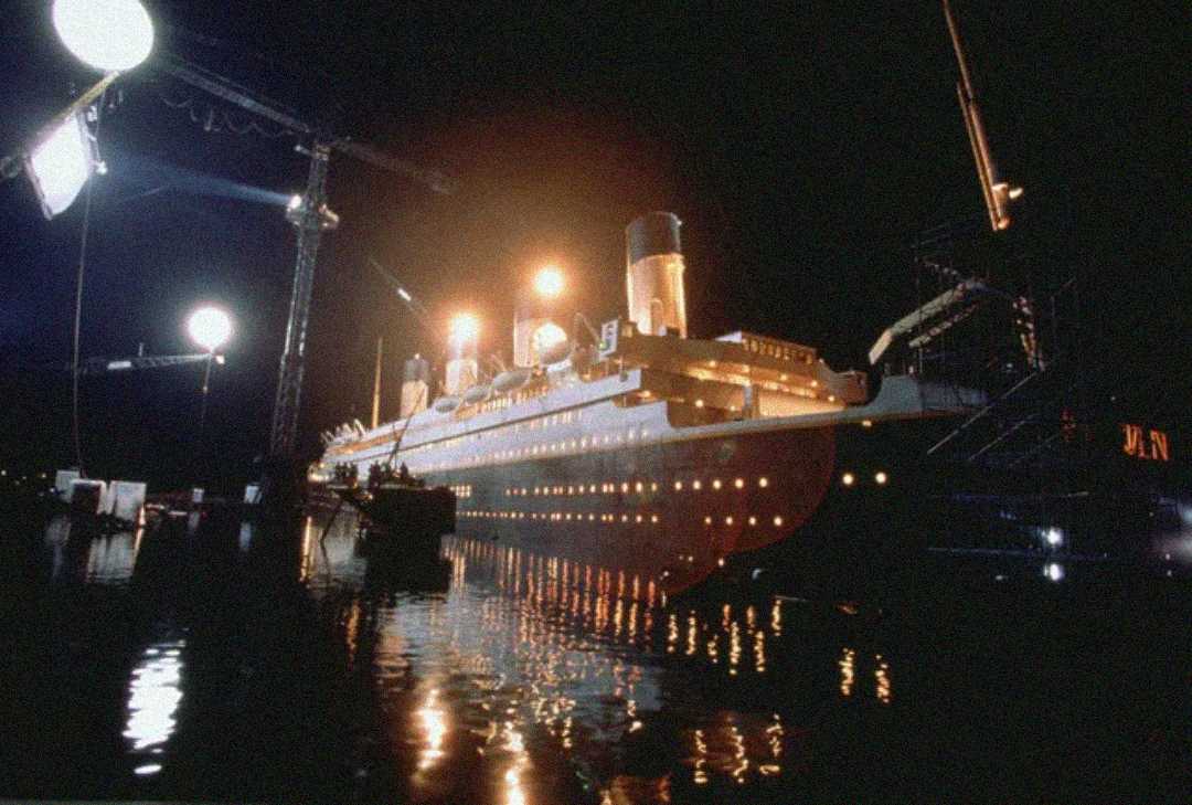 A large film set at night features a replica of an ocean liner, resembling the Titanic, illuminated by bright studio lights with cranes and crew visible around water at the base.