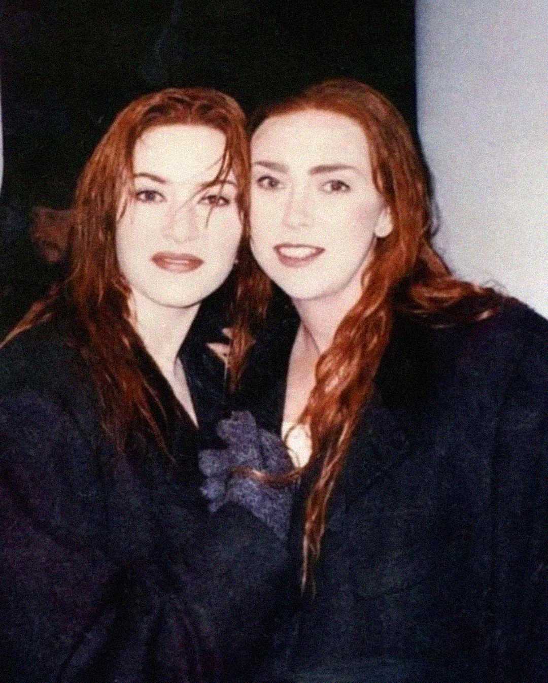 Two women with long auburn hair, wearing dark coats, stand close together and smile at the camera. The background is dark, and their hair appears wet.
