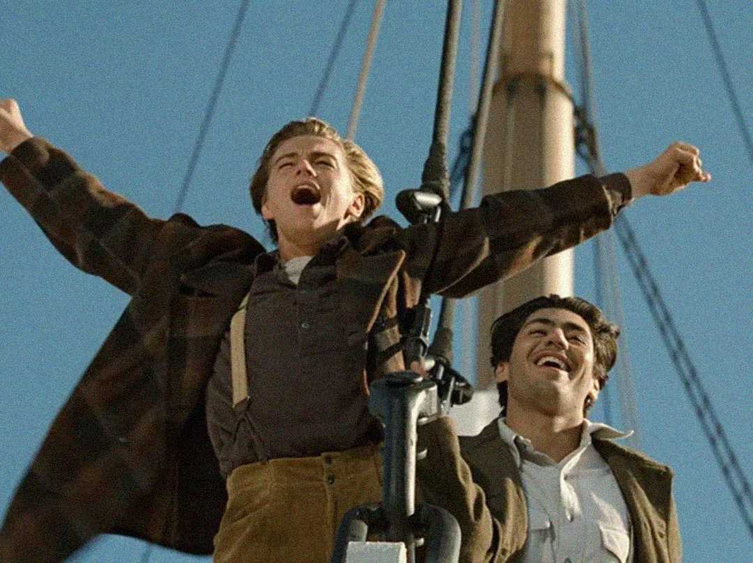 Two young men stand at the bow of a ship with arms outstretched, one with eyes closed and mouth open in excitement, recreating the famous scene from the movie Titanic against a clear blue sky.