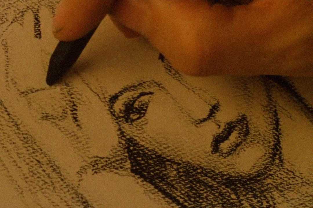 A close-up of a hand drawing a woman's face with a black pastel on textured paper. The sketch shows the woman's eyes, nose, and lips, and the artist's fingers hold the pastel crayon above the drawing.