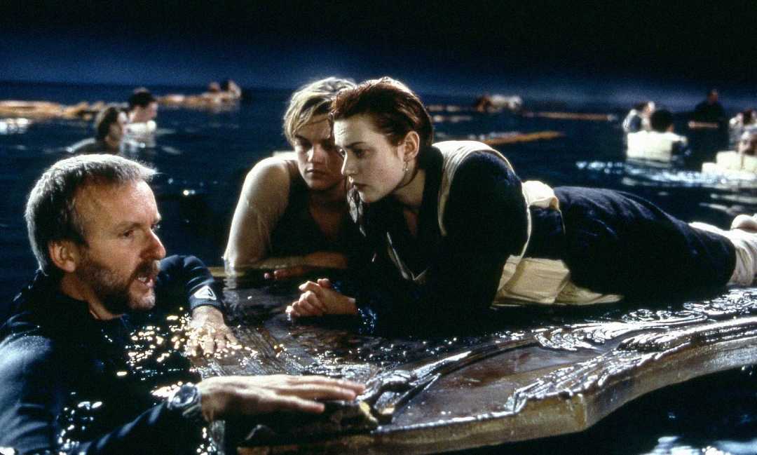A film director speaks to two actors on a large wooden panel in water, recreating a famous Titanic scene. Other extras float in the background, simulating a nighttime ocean disaster.