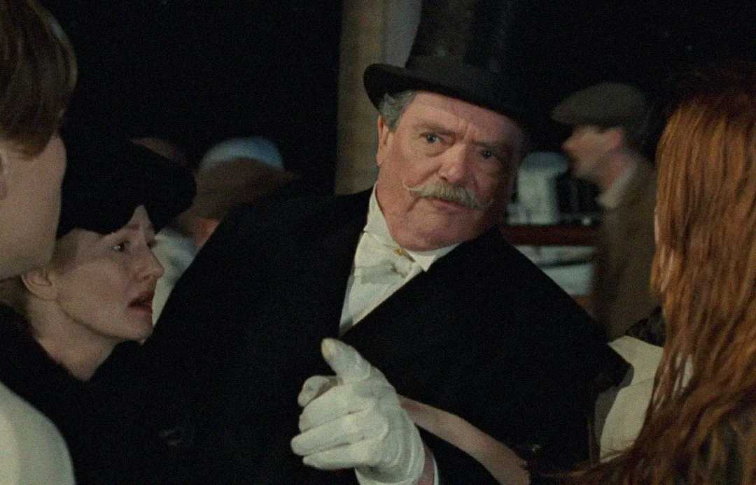 An older man with a mustache, wearing a black coat, white gloves, and a bowler hat, points forward while surrounded by concerned people in period clothing. The setting appears to be from an early 20th-century era.