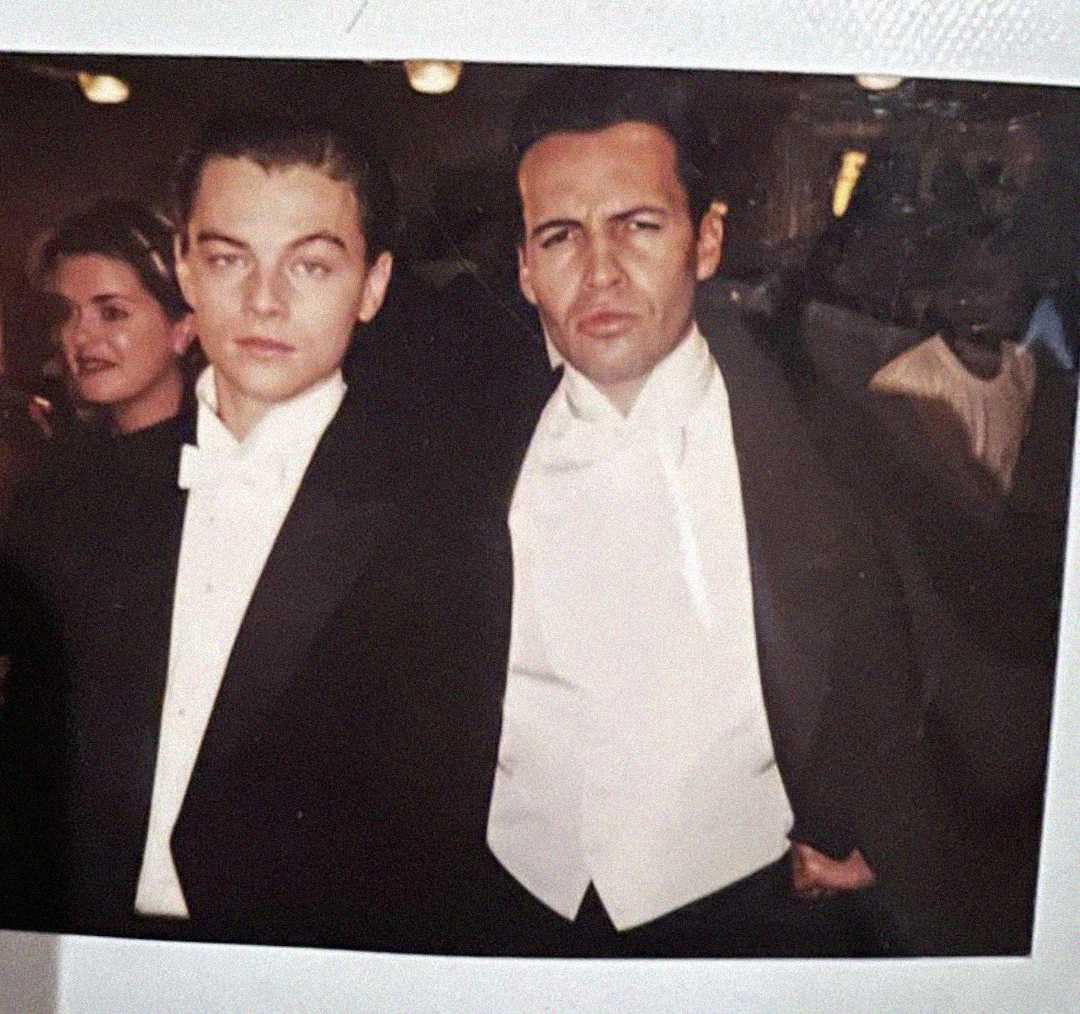 Two men in formal black tuxedos and white bow ties pose together at an event; one has blond hair, the other dark hair. A woman stands in the background, also dressed formally.