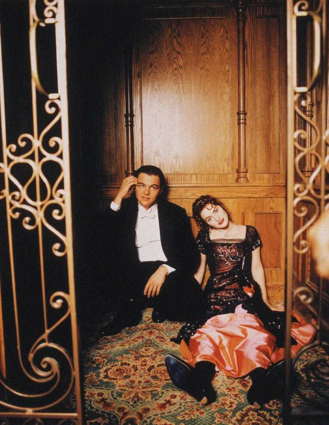 A man in a tuxedo and a woman in an elegant red and black dress sit on a patterned carpeted floor, leaning against wooden paneling, seen through ornate iron gates. The woman looks relaxed with her head tilted.