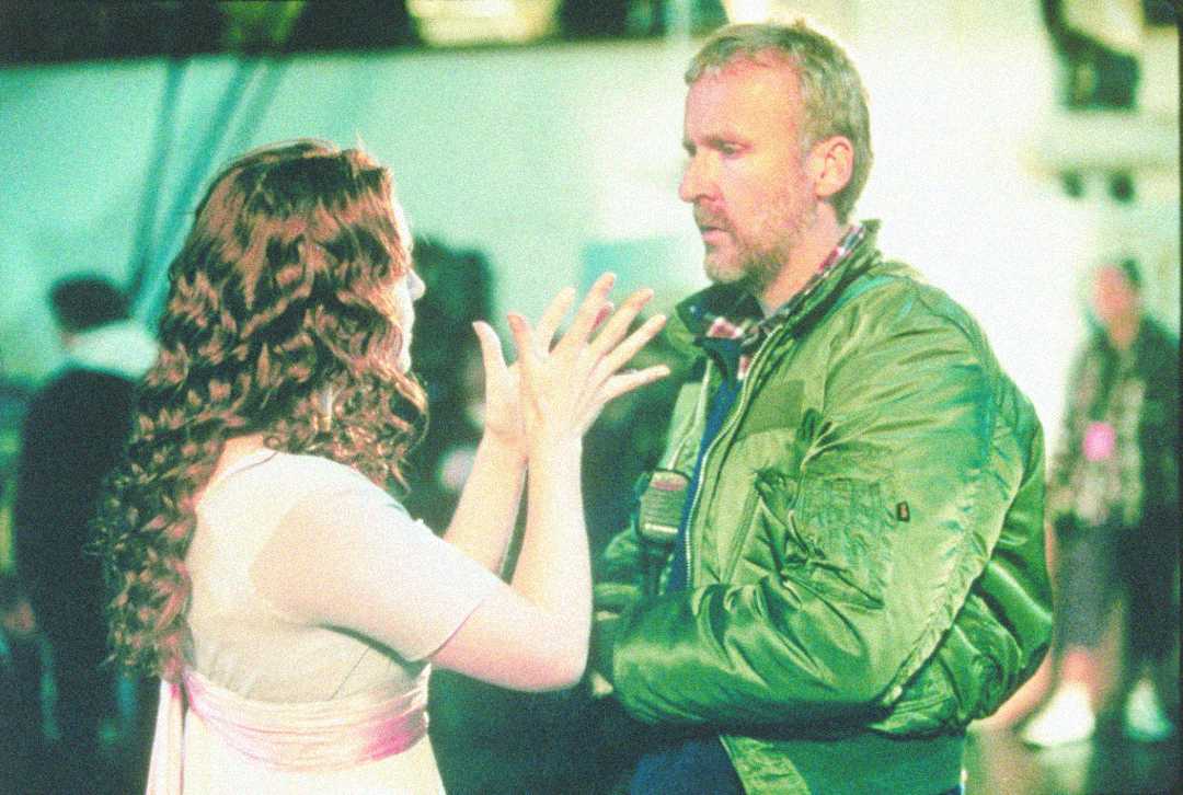 A woman in a light dress gestures with her hands while facing a man in a green jacket, who is listening intently on a film set with blurred people and equipment in the background.