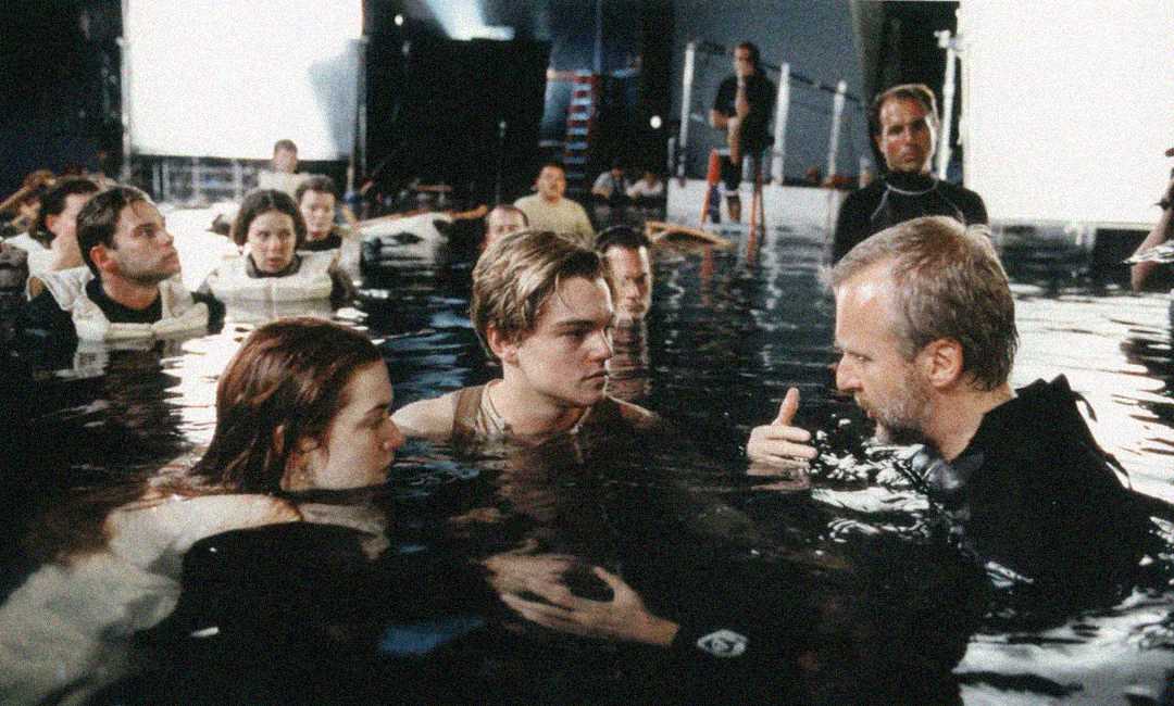 Actors and crew stand in waist-deep water on a film set, as a director gives instructions to two main actors who are facing him, surrounded by lights and equipment.