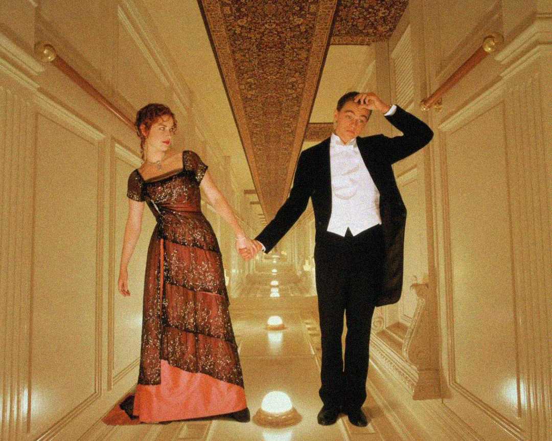 A woman in a red and black dress and a man in a black tuxedo hold hands while standing in a hallway, with the image rotated to make it appear as if they are standing on the wall.