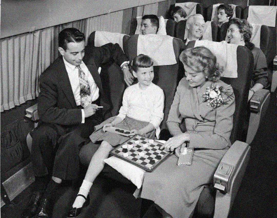 A black-and-white photo of three people in an airplane row: a man, a young girl, and a woman playing checkers. Other passengers sit behind them, appearing relaxed and engaged in conversation.