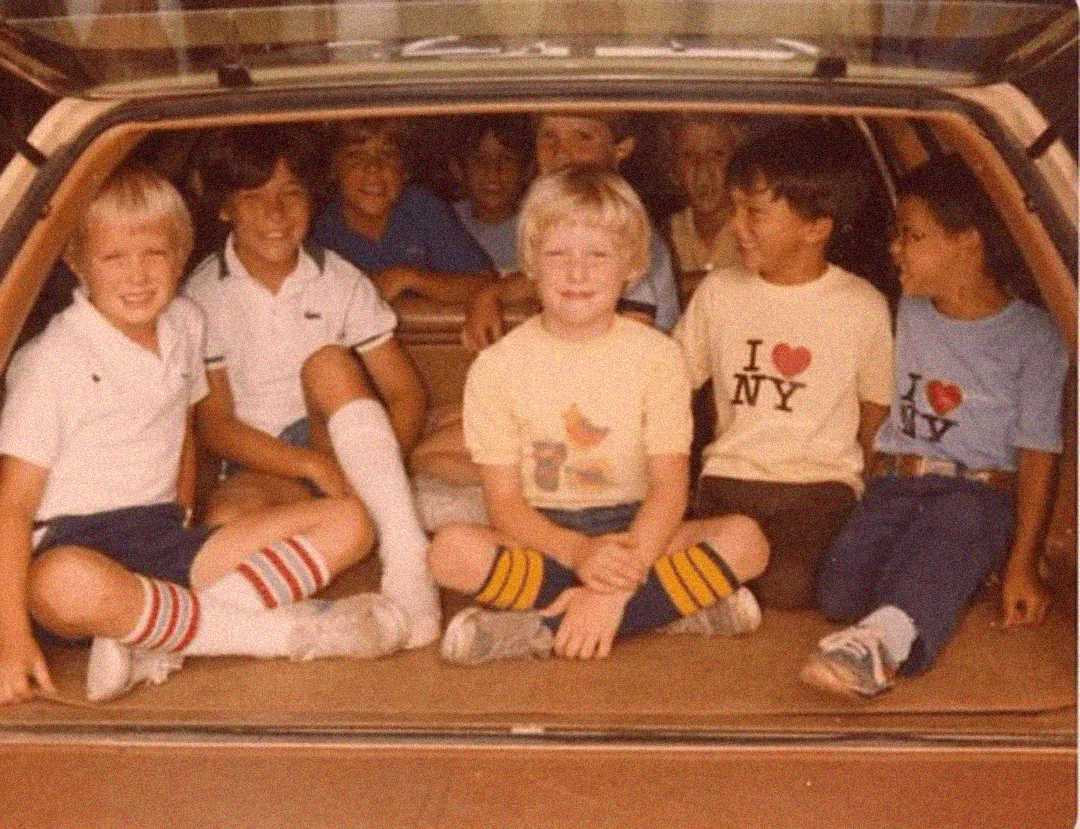 A group of eight children sits together in the back of a station wagon. They are smiling and wearing casual 1970s or 1980s clothing, including striped socks and "I ♥ NY" T-shirts.