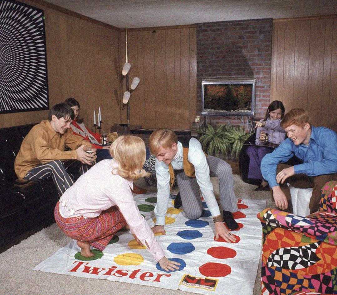 Five teenagers are in a wood-paneled living room. Three play Twister on the floor while two sit on a couch watching. The room is decorated with retro furniture, a patterned blanket, and a brick fireplace.