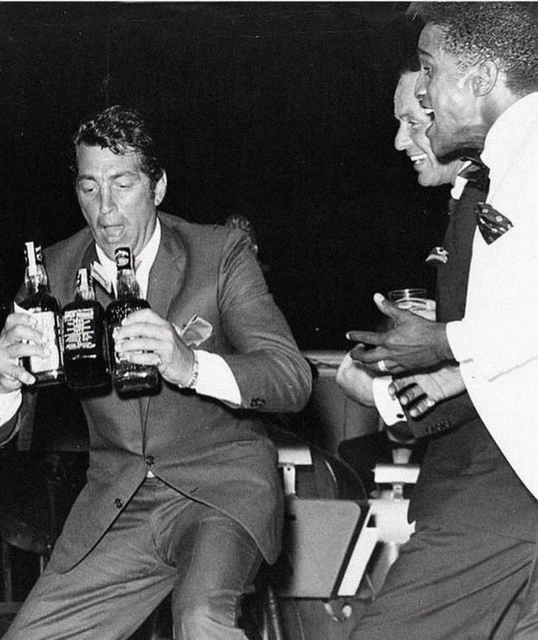 Three men in suits are laughing and interacting; one man holds three bottles of liquor while the others watch and smile, creating a lively and joyful atmosphere. The image is in black and white.