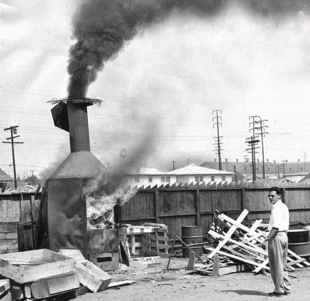 A man in a white shirt and sunglasses stands near a smoking incinerator releasing thick black smoke in a yard filled with wooden debris and pallets, with power lines and buildings visible in the background.