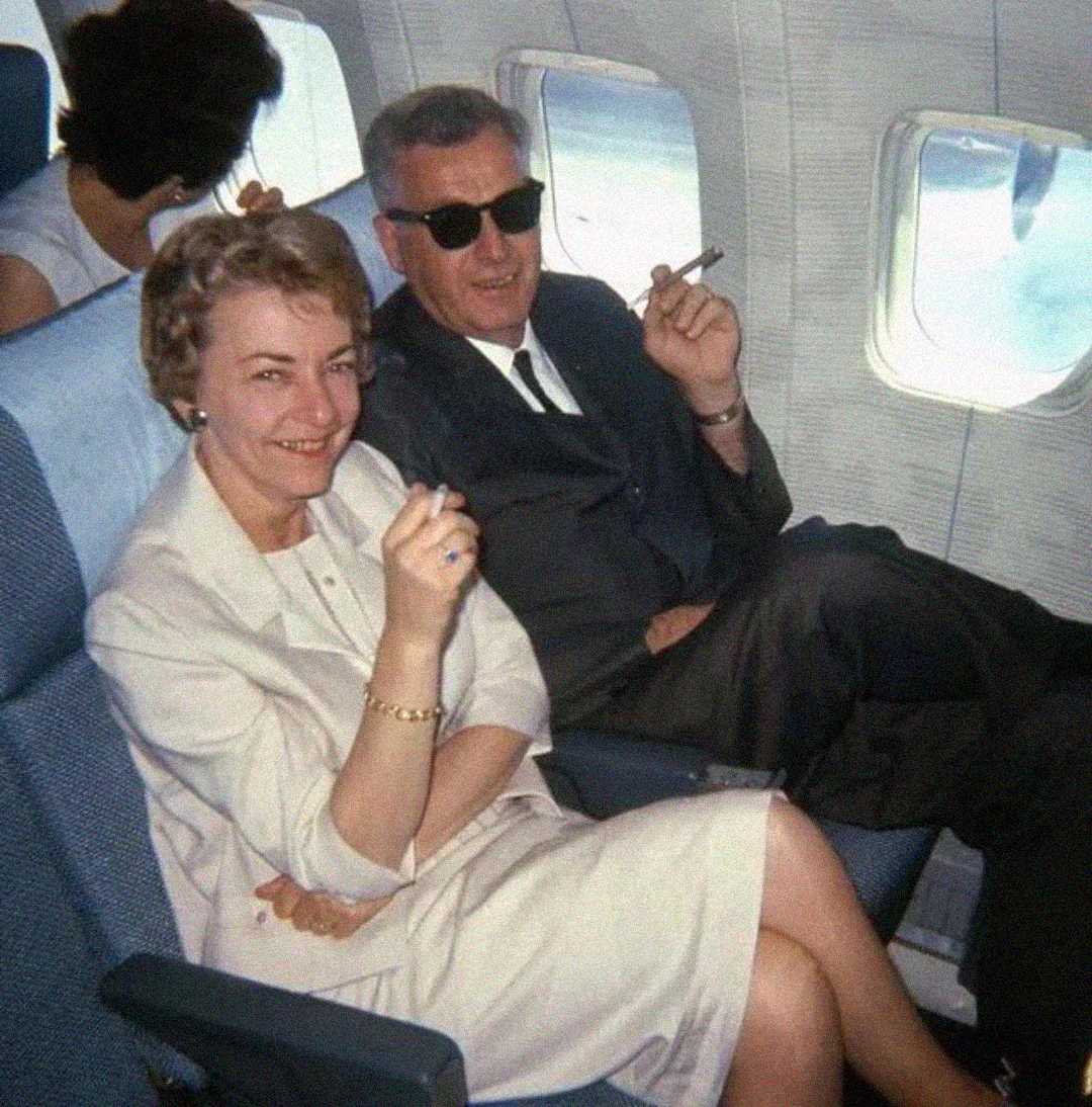 A man and a woman sit side by side on an airplane, both smiling and holding cigars. The man wears a suit and sunglasses, while the woman wears a light-colored outfit. Another passenger sits behind them.