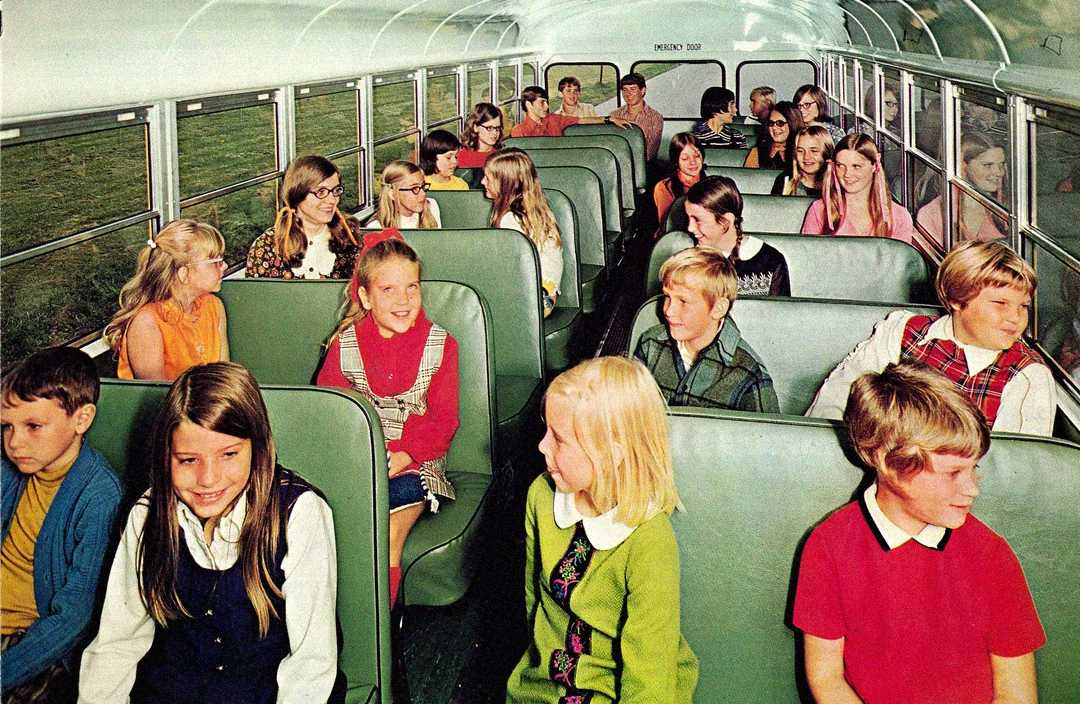 Children sit on green bench seats inside a school bus, chatting and smiling. The bus is filled with natural light from large windows, and the kids wear colorful, vintage-style clothing.