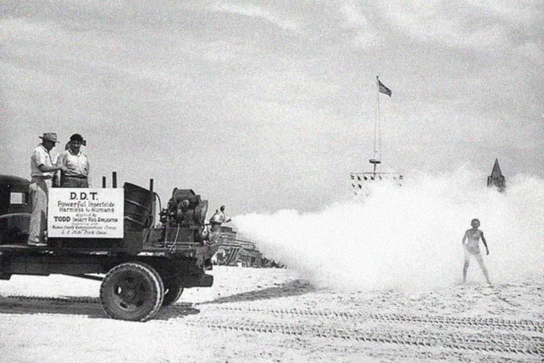 A black-and-white photo shows a truck spraying a large cloud of DDT insecticide on a beach, with people nearby and an American flag in the background. A few individuals watch from the truck.