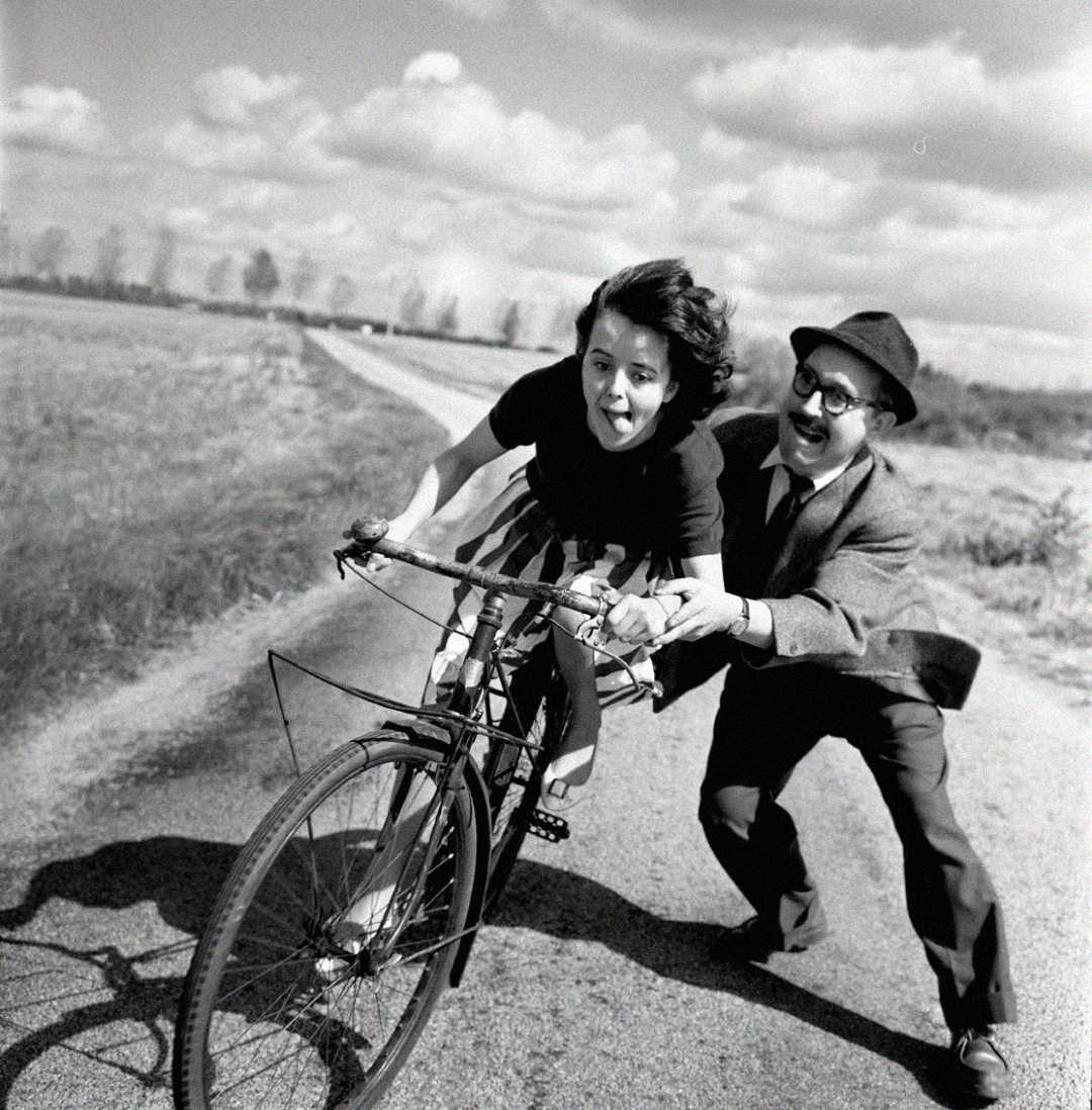 A man in a suit and hat helps push a smiling woman riding a bicycle on a rural road, with fields and a cloudy sky in the background.