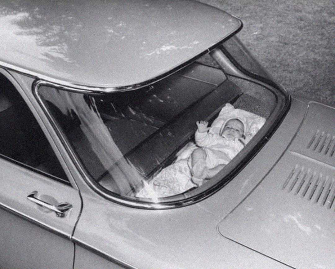 A black and white photo of a baby lying on a blanket in the rear window shelf of a classic car, seen from above. The baby is looking up and waving, with trees and grass visible outside.
