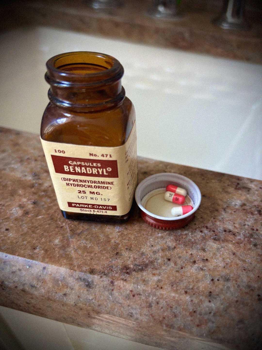 An open brown glass bottle of Benadryl capsules sits on a bathroom counter. The cap, placed beside it, contains three pink and white capsules. The label on the bottle is clearly visible.