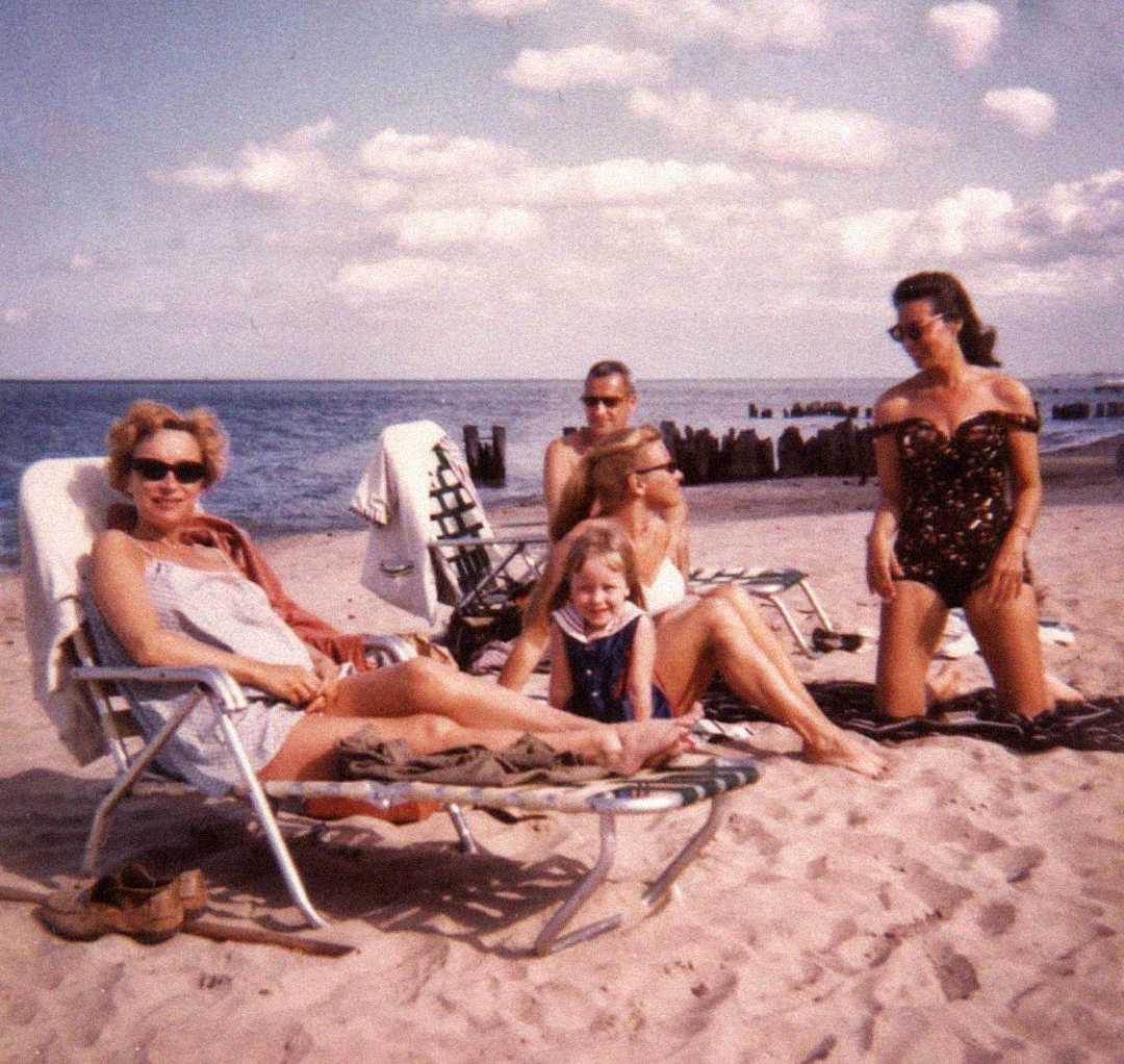 Four adults and a child relax on a sandy beach. Two women sit on lounge chairs, one man sits beside them, and another woman stands nearby in a black swimsuit. The ocean and scattered clouds are in the background.