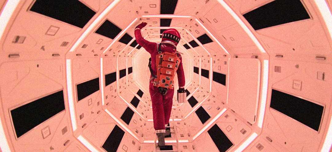 An astronaut in a red spacesuit walks through a brightly lit, hexagonal spaceship corridor filled with geometric patterns and panels.