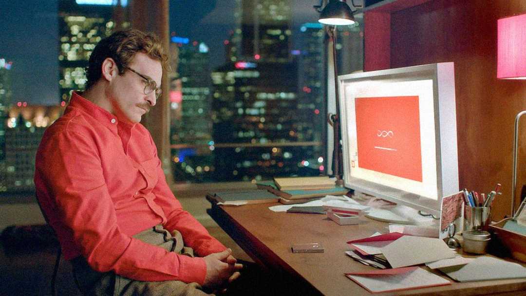 A man in glasses and a red shirt sits at a desk in a dimly lit office, looking thoughtfully at a computer screen with a red background. The city skyline is visible through the window behind him.