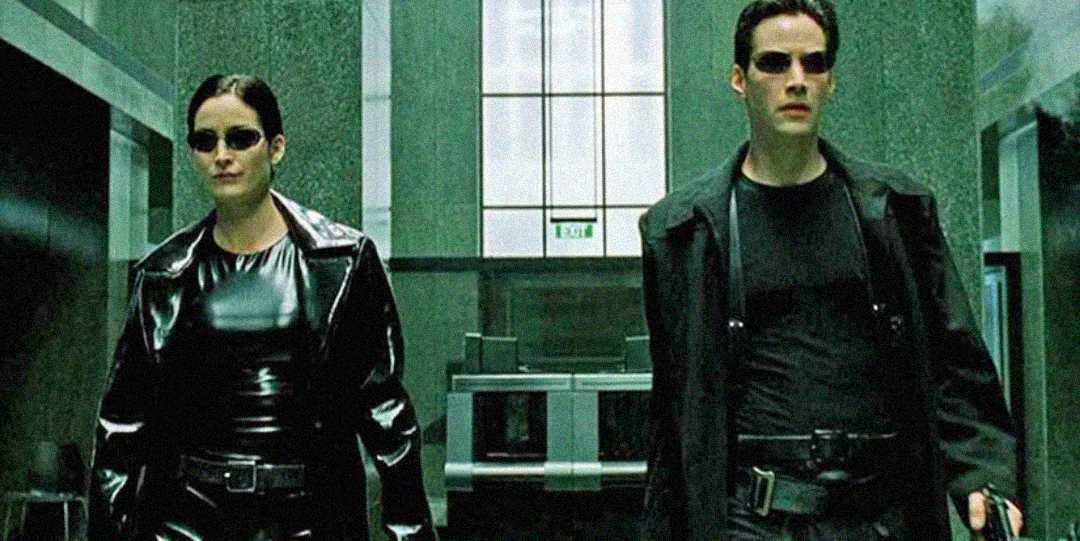 Two people in black leather outfits and sunglasses walk confidently through a green-tinted, modern building corridor. Their serious expressions and attire give a futuristic, action-movie vibe.