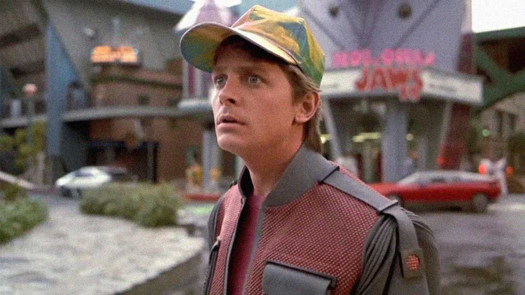 A young man wearing a colorful, reflective cap and a futuristic jacket stands outdoors, looking surprised. The background features modern buildings and a car driving by.