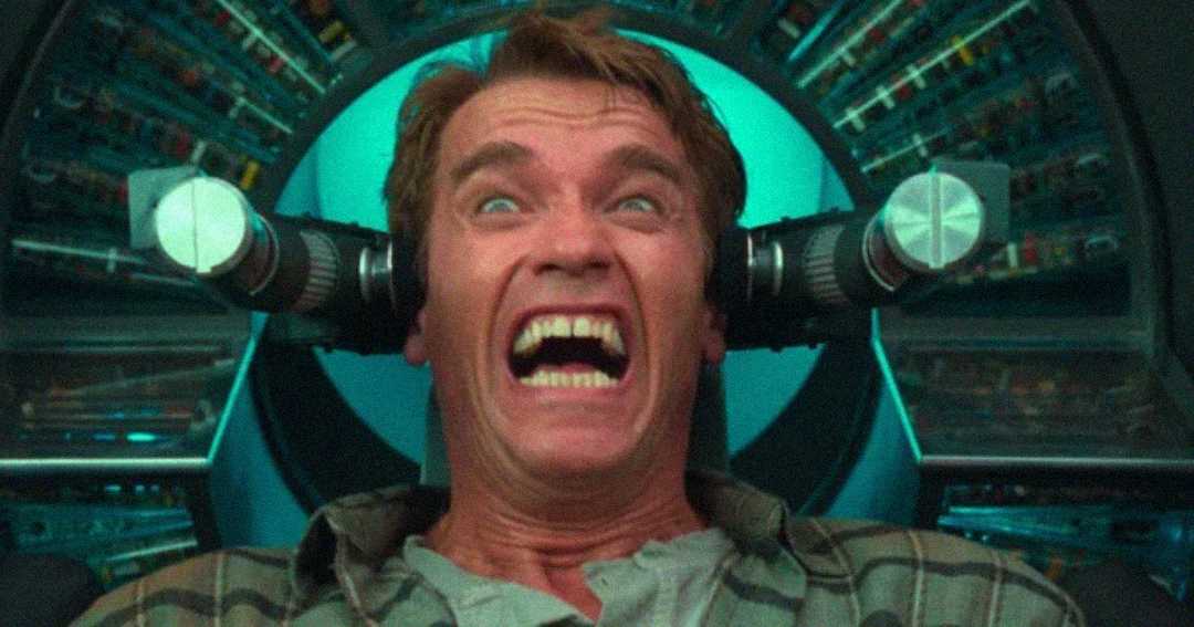 A man with a fearful, wide-eyed expression sits in a high-tech chair with metal devices positioned near his head, as if undergoing a procedure or experiment.