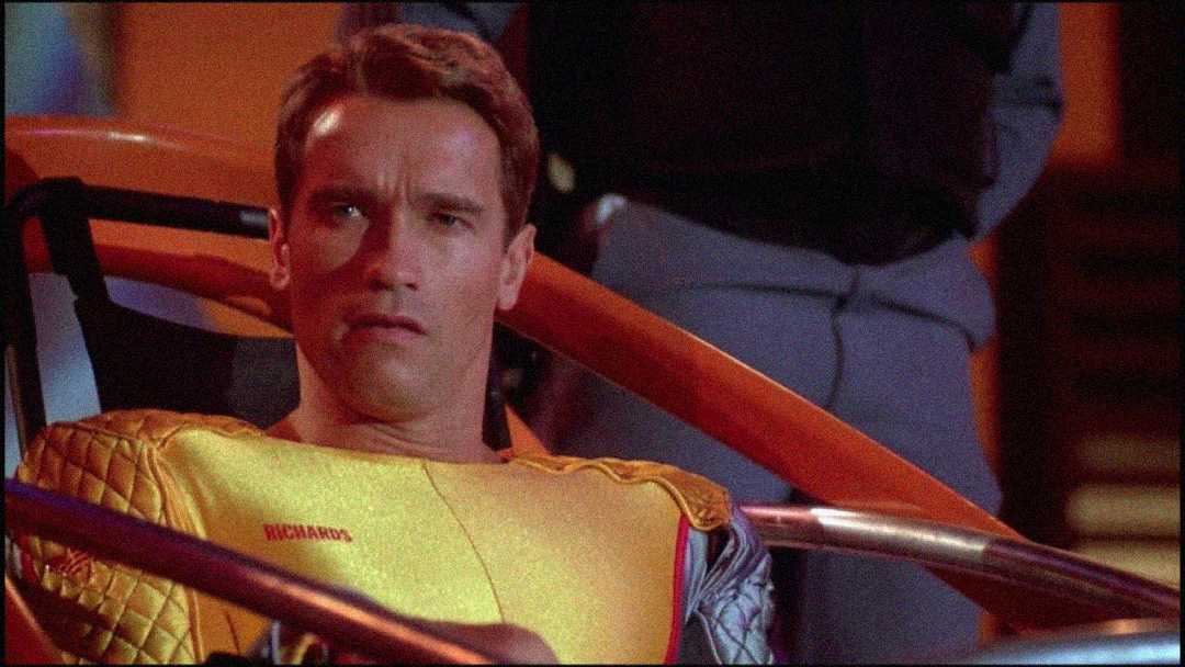 A man with short hair wearing a yellow and silver futuristic suit sits in a vehicle, looking forward with a serious expression. The background is out of focus and features orange and grey colors.