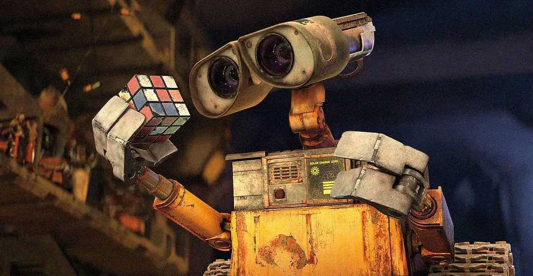 A rusty, boxy robot with large binocular eyes holds a colorful Rubik's Cube in one hand, studying it intently. The robot appears curious, with gears and metal parts visible in a dimly lit, cluttered background.