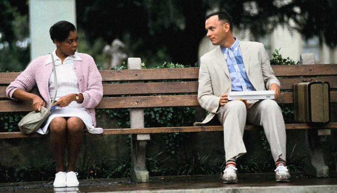 A man in a light suit sits on a park bench holding a box of chocolates, looking at a woman in a pink sweater who is seated at the other end of the bench, with trees and greenery in the background.