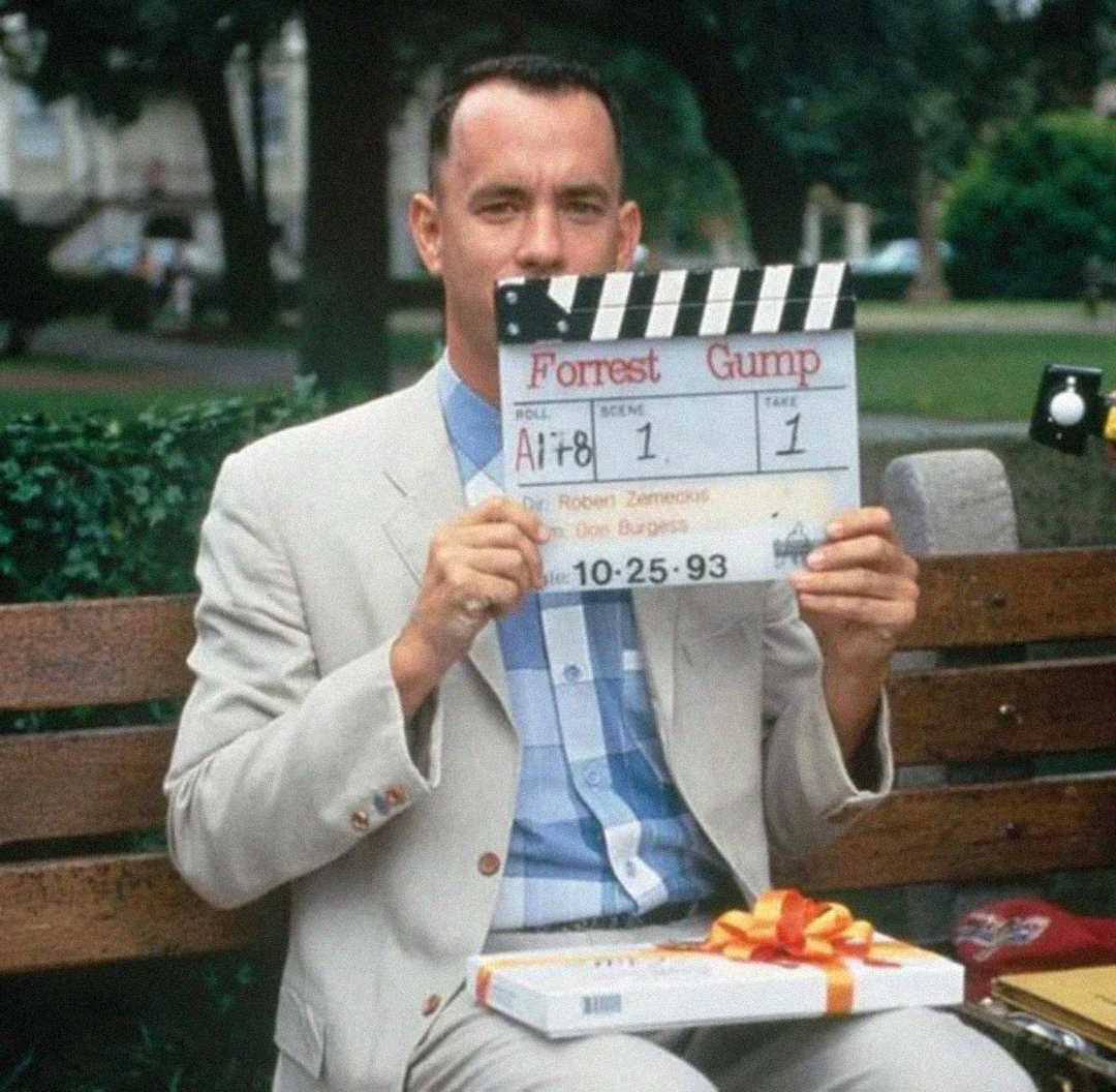 A man in a light suit sits on a bench, holding a clapperboard labeled "Forrest Gump" in front of his face. A box of chocolates with a ribbon rests on his lap. Trees and grass are in the background.