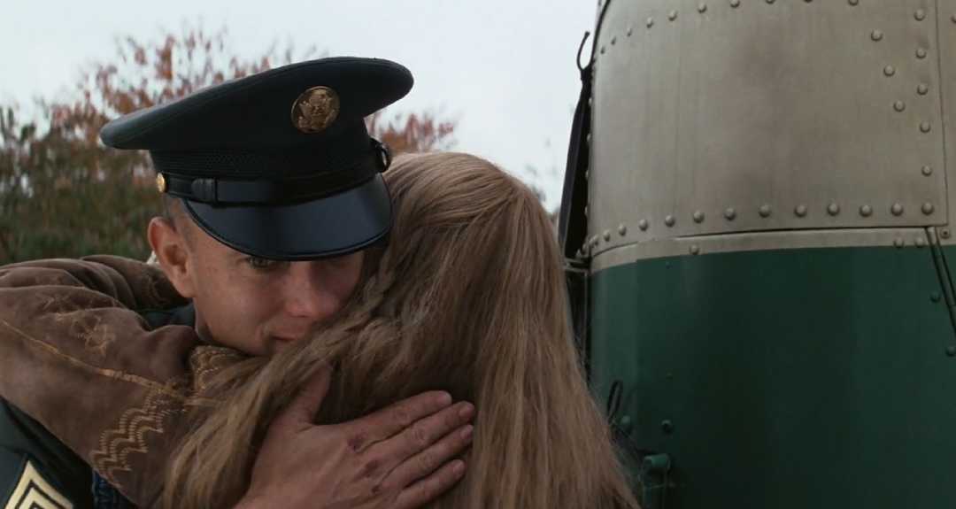 A man in a military uniform hugs a woman with long blonde hair tightly near a green and silver bus, both appearing emotional. Trees with autumn leaves are visible in the background.