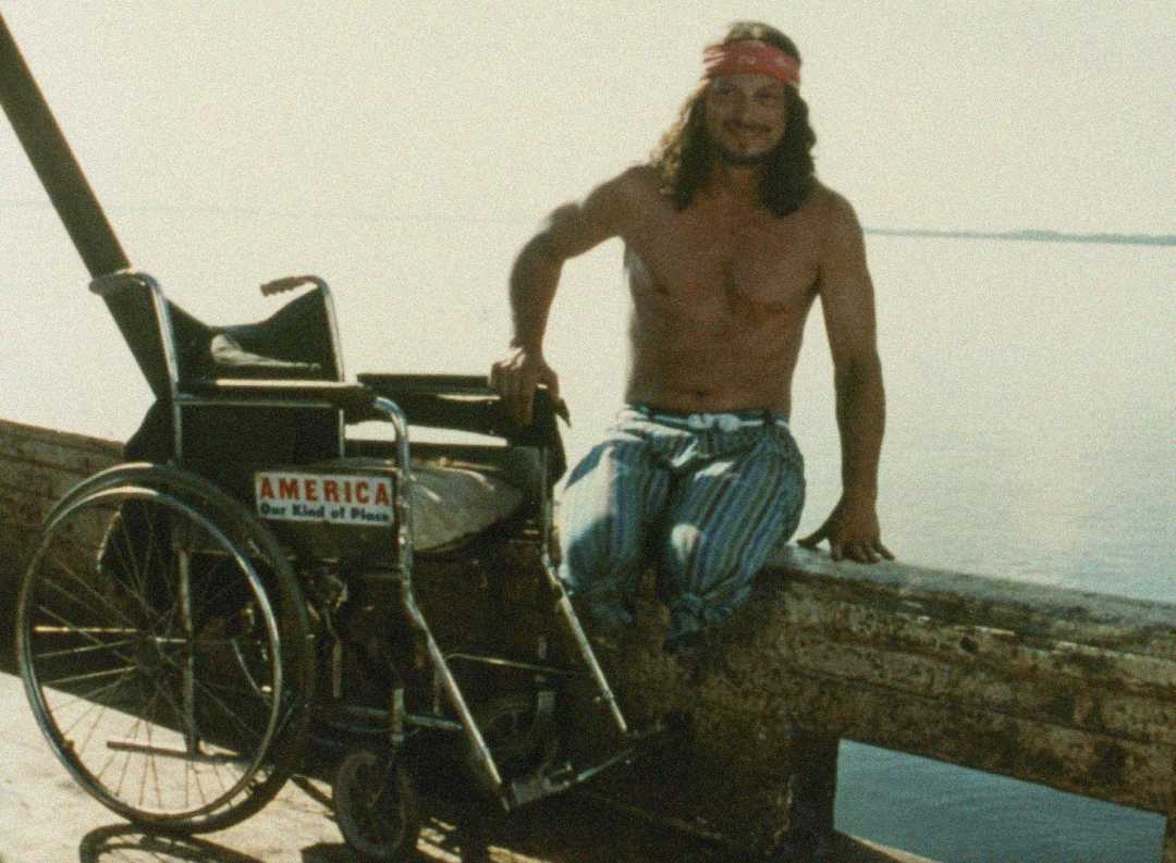 A shirtless man with long hair and a red headband sits on a wooden railing by the water next to a wheelchair with a sticker that reads "AMERICA Our Kind of Place.