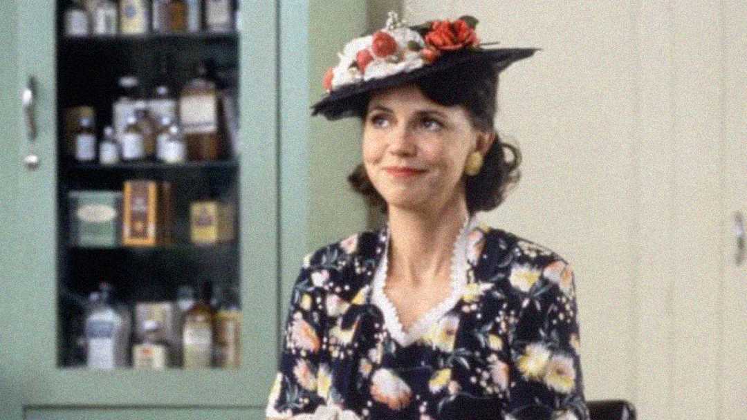 A woman wearing a floral dress and a decorative hat with flowers sits indoors, smiling. Behind her is a green cabinet with shelves holding various bottles and jars.