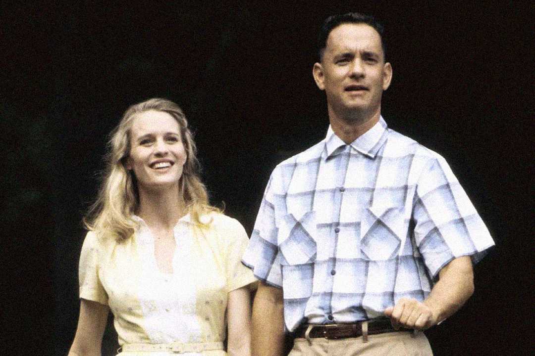 A woman in a light yellow dress and a man in a short-sleeved plaid shirt stand side by side outdoors, both looking ahead; the man holds his right arm bent as if mid-walk.